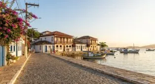 Caminho de pedra à beira-mar com casas coloridas e barcos ao fundo, flores na calçada e luz do sol ao entardecer.
