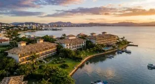 Vista aérea de hotéis à beira-mar em Cacupé, com montanhas ao fundo e barcos ancorados.