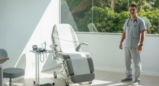 Médico proctologista em consultório moderno, com cadeira de exame e equipamentos de saúde ao fundo.