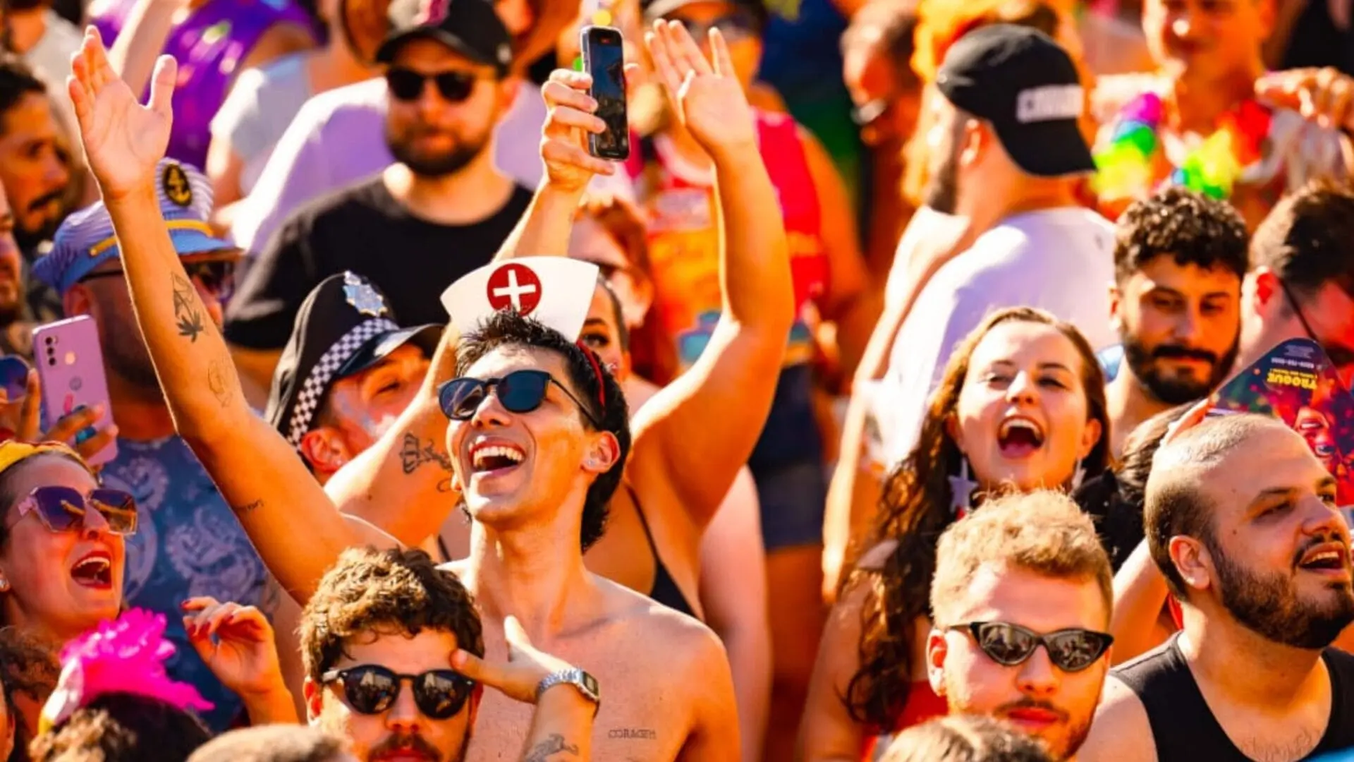 Multidão animada durante o Carnaval, pessoas sorrindo, com fantasias coloridas e mãos levantadas.