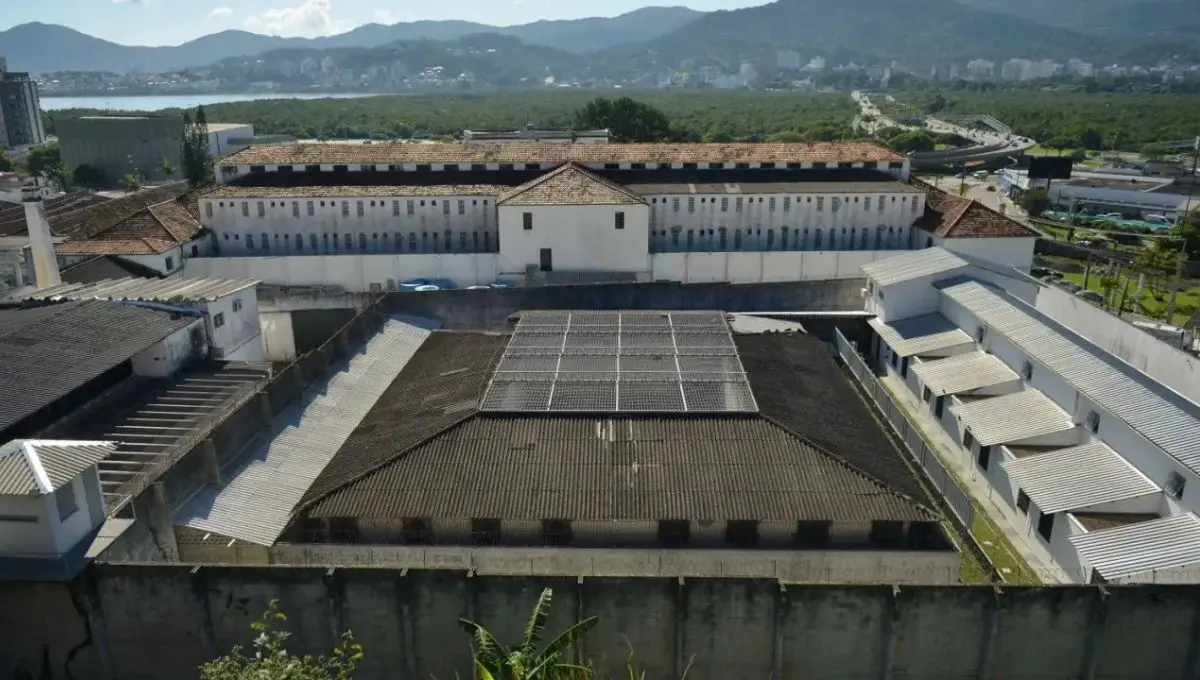 Vista aérea de um complexo prisional com pátio central, cercado por muros e montanhas ao fundo.