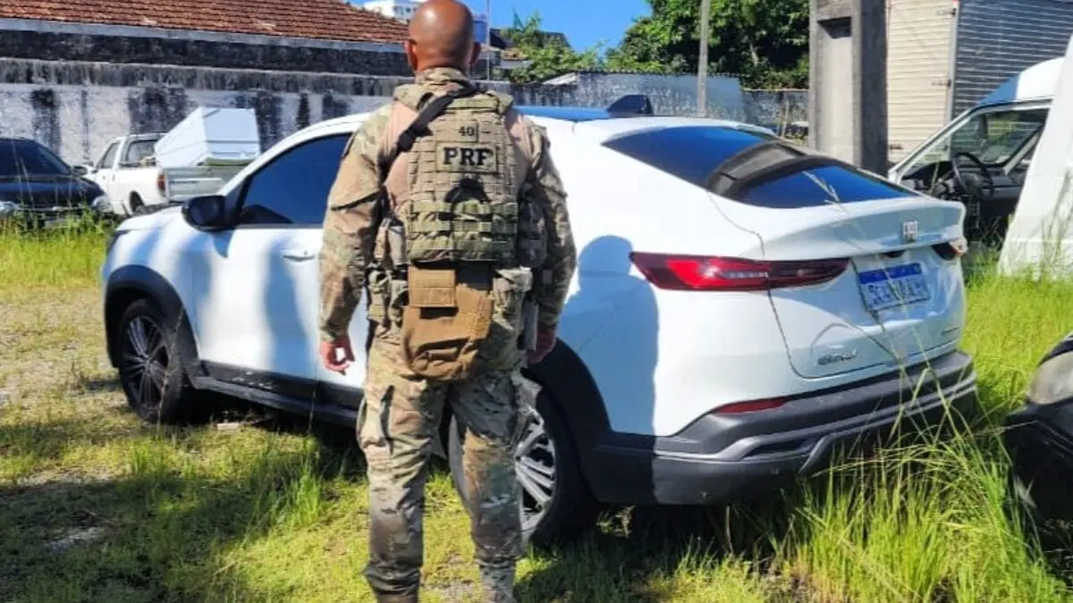 Policial da PRF observa carro branco estacionado em área com grama alta, em operação de fiscalização.