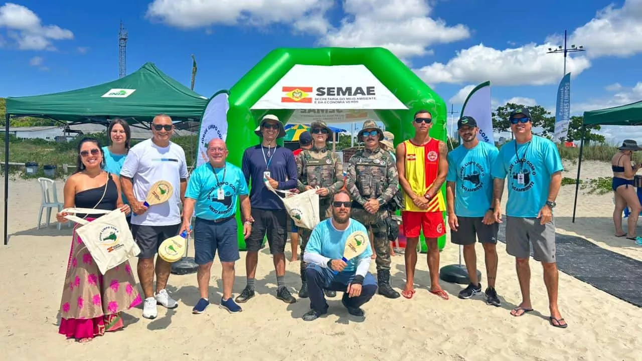 Grupo de pessoas em frente a um arco verde, com camisetas azuis, na praia de Canasvieiras, sob céu azul e nuvens.