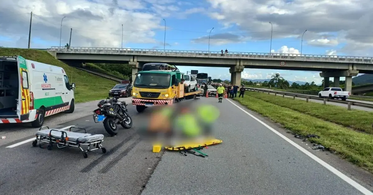 Acidente na BR-101 com veículos parados, ambulância, moto e equipe de resgate na pista. Céu nublado ao fundo.