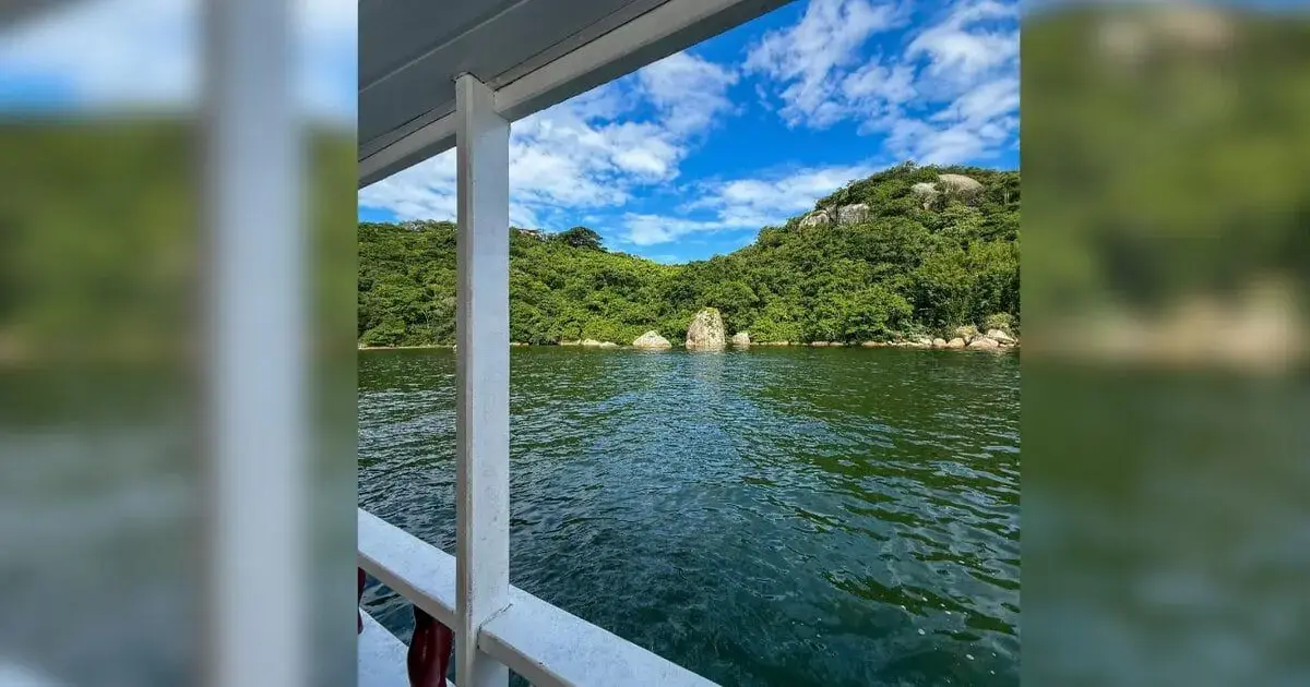 Vista de um lago calmo com vegetação ao fundo e céu azul com nuvens.