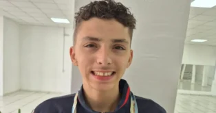 Adolescente sorridente com cabelo cacheado, usando jaqueta escura, em ambiente interno com piso claro.