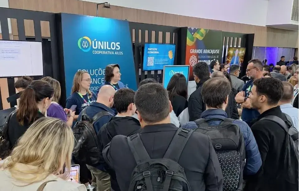 Grupo de pessoas em evento, interagindo em estande da Ailos Únilos, com painéis informativos ao fundo.