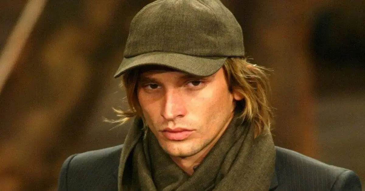 Homem com cabelo longo, usando boné e cachecol, com expressão séria em ambiente interno.