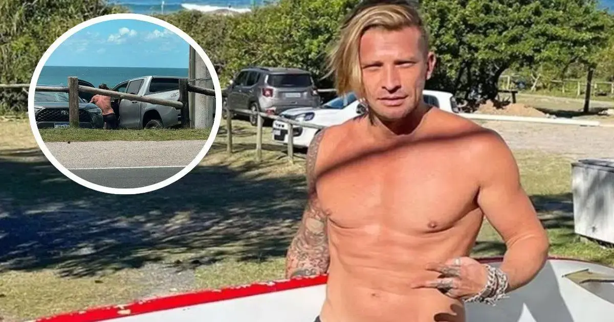 Homem sem camisa posando em área externa, ao fundo, veículo estacionado e pessoas ao longe.