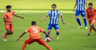 Jogadores em campo durante partida de futebol, um atleta com camisa azul e branca driblando adversários em laranja.