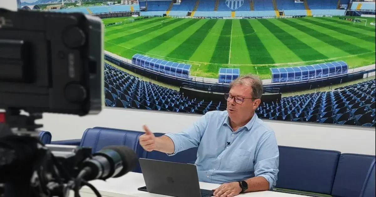 Homem de camisa clara gesticulando em frente a um computador, com campo de futebol ao fundo.
