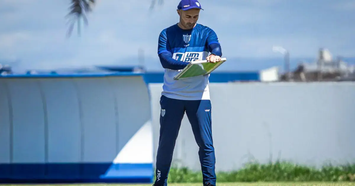 Treinador do Avaí analisando táticas em campo, vestindo uniforme azul e branco, sob céu claro.
