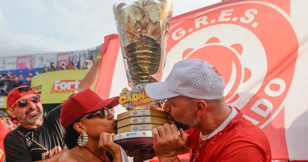 Casal se beijando em frente ao troféu do bicampeonato, com bandeira vermelha ao fundo e pessoas sorrindo.