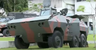 Blindado anfíbio camuflado em tons de verde e marrom, estacionado em área externa com vegetação ao fundo.