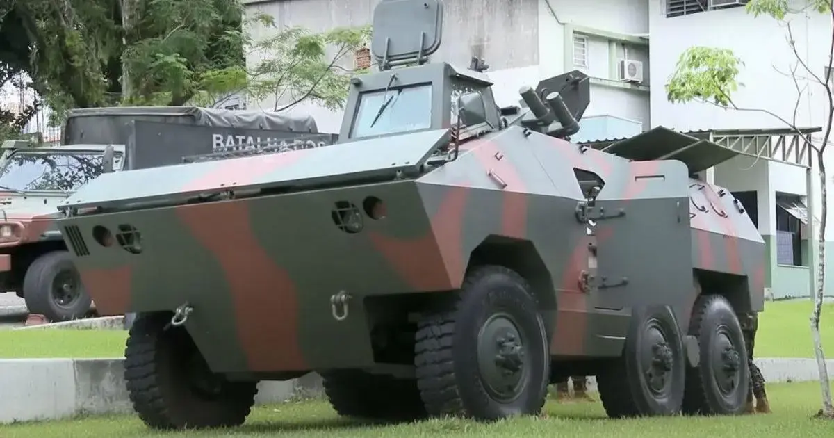 Blindado anfíbio camuflado em tons de verde e marrom, estacionado em área externa com vegetação ao fundo.