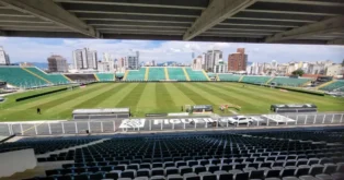 Estádio com gramado bem cuidado, arquibancadas vazias e prédios ao fundo em dia ensolarado.
