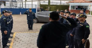 Grupo de homens em uniforme militar, alguns em posição de saudação, em ambiente externo com veículos e estrutura ao fundo.