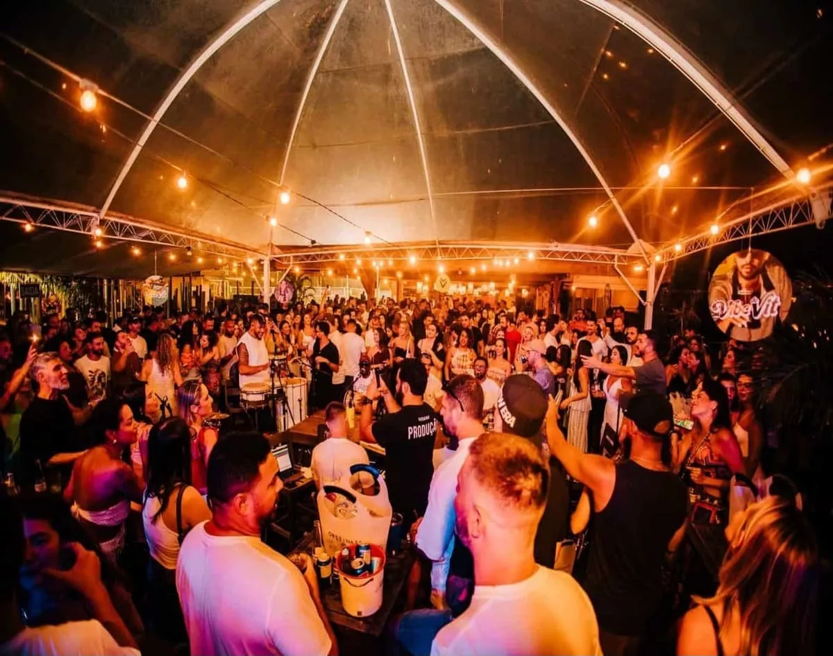 Multidão animada em festa sob tenda iluminada, com pessoas dançando e se divertindo em ambiente festivo.