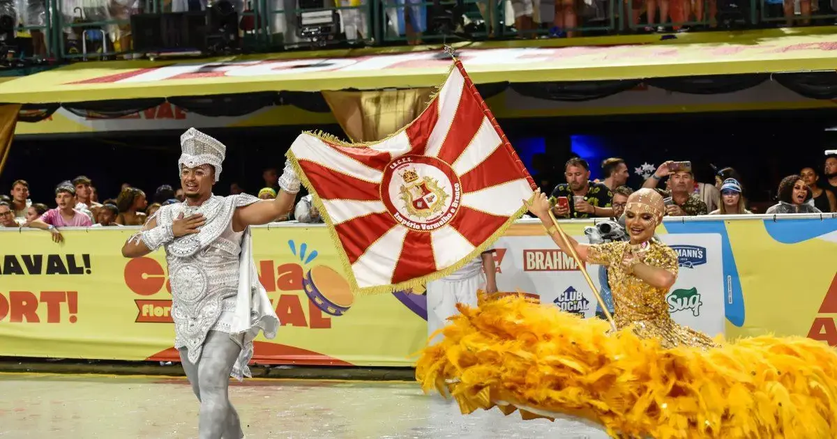 Dançarinos de carnaval desfilam com bandeira e plumas amarelas em ambiente festivo com público ao fundo.