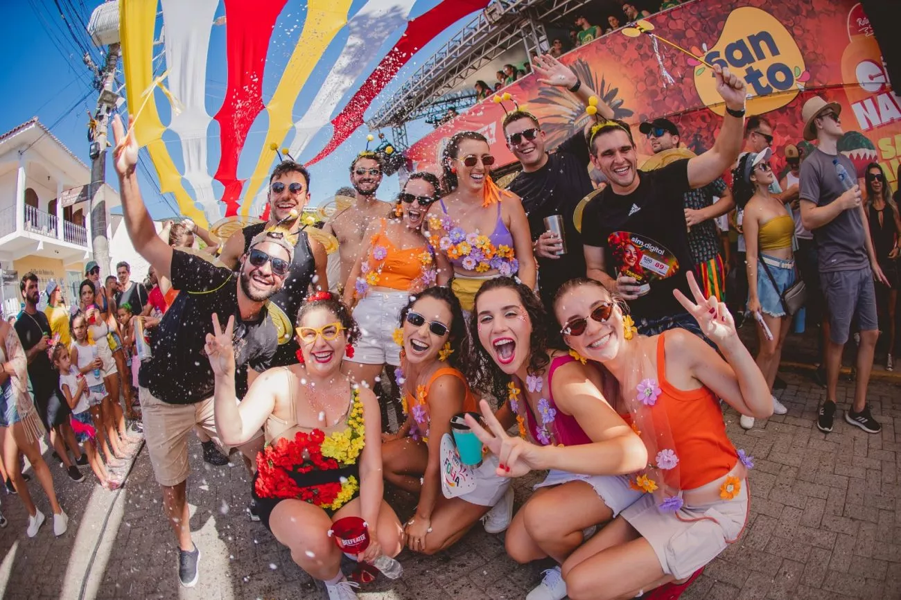 Grupo de pessoas sorrindo e se divertindo em um carnaval de rua, com roupas coloridas e adereços festivos.