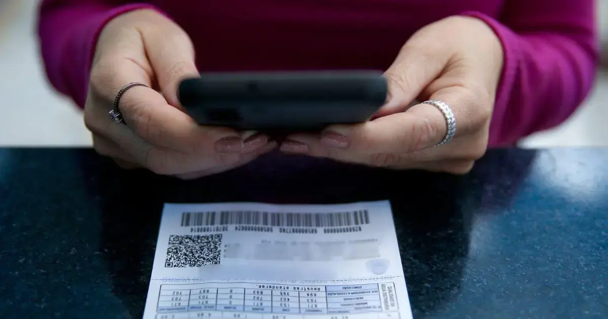 Mãos segurando um celular acima de uma conta de água em uma superfície escura.