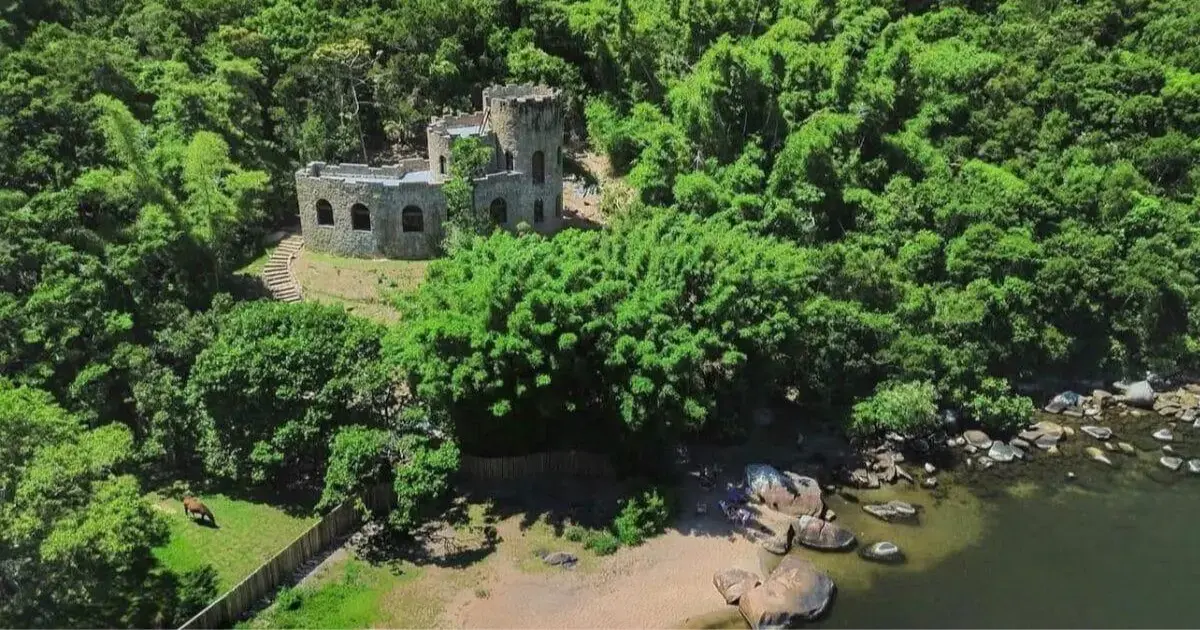 Castelo em ruínas cercado por vegetação densa, próximo à praia e rochas à beira-mar.