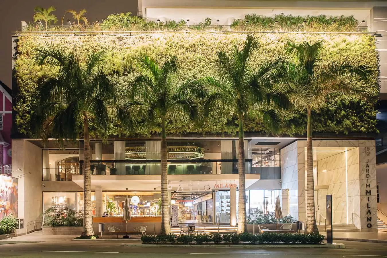 Fachada de prédio com jardim vertical e palmeiras em frente, iluminado à noite.