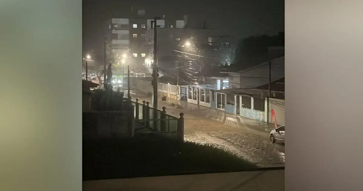 Rua alagada à noite, com postes de luz acesos e prédios ao fundo, indicando forte chuva na Grande Florianópolis.