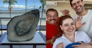 Bolo gigante em formato de pedra, confeiteiros sorrindo ao fundo, céu azul e palmeiras visíveis.