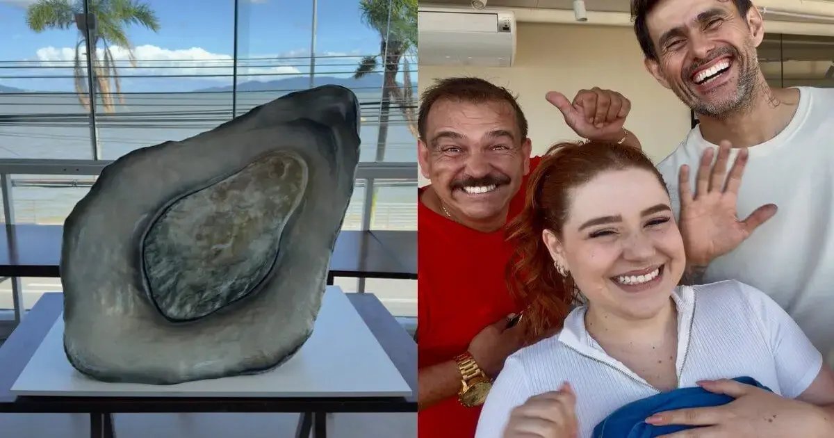 Bolo gigante em formato de pedra, confeiteiros sorrindo ao fundo, céu azul e palmeiras visíveis.