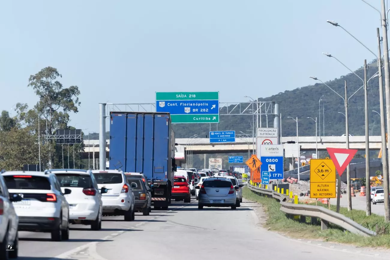 Trânsito intenso na rodovia com placas indicativas e caminhões, ao fundo montanhas e céu claro.