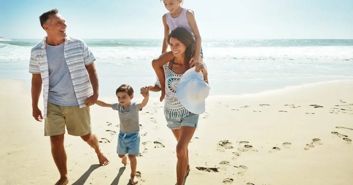 Família caminhando na praia, crianças brincando, sol brilhante e mar ao fundo.