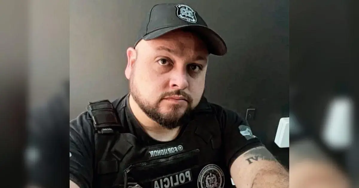 Homem com barba e boné, usando colete tático e camisa preta, posando para a câmera em ambiente interno.