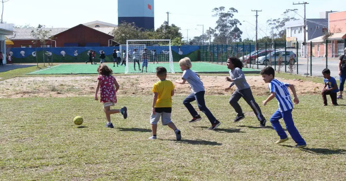 Crianças jogando futebol em campo, algumas correndo atrás da bola, outras observando.