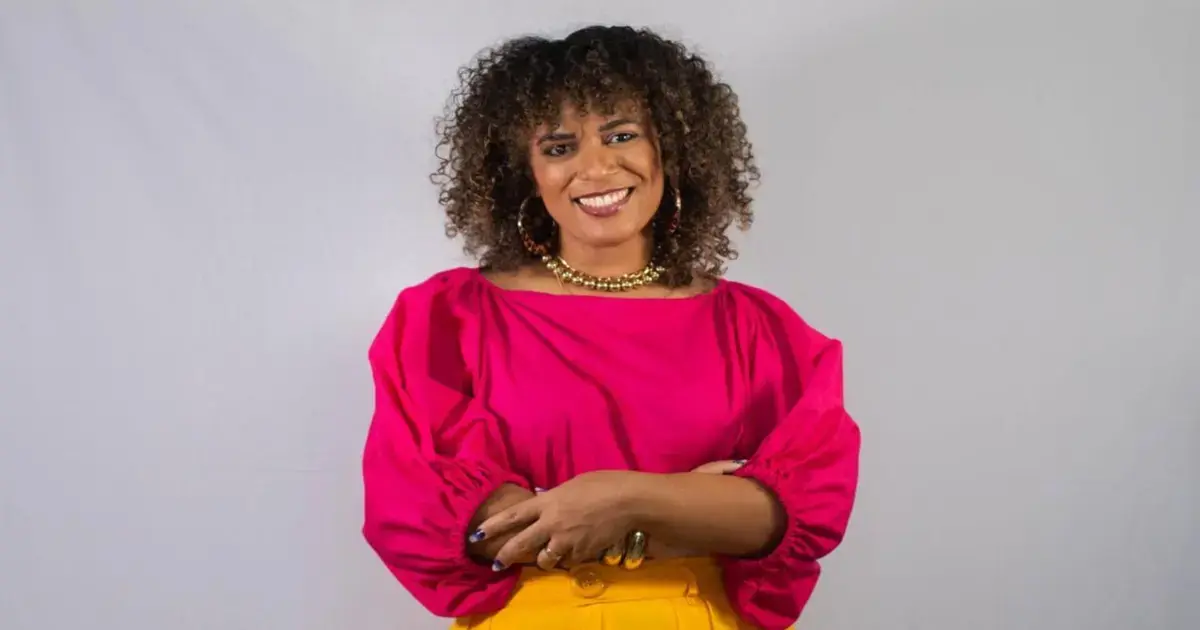 Mulher sorridente com cabelo cacheado, usando blusa rosa e saia amarela, posando com os braços cruzados.