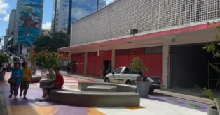 Calçadão com bancos e árvores, lojas fechadas ao fundo, pessoas caminhando e mural colorido na parede.