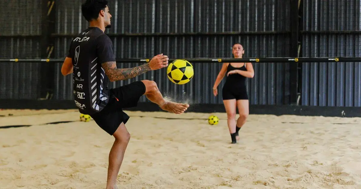 Homem com tatuagens jogando futevôlei na areia, enquanto mulher observa ao fundo, ambos em trajes de banho.