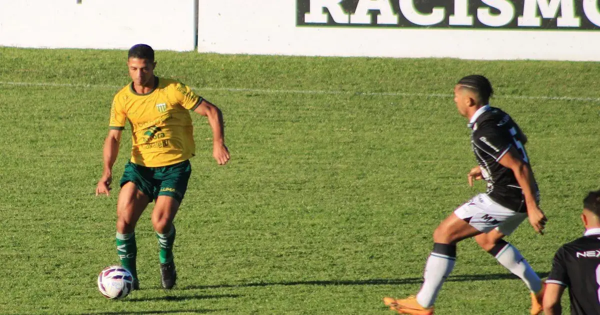 Jogadores em campo durante partida de futebol, um com uniforme amarelo e verde, outro com uniforme preto e branco.