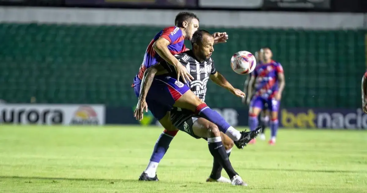 Jogadores em campo disputando a bola, um atleta em pé e outro caindo, gramado ao fundo.