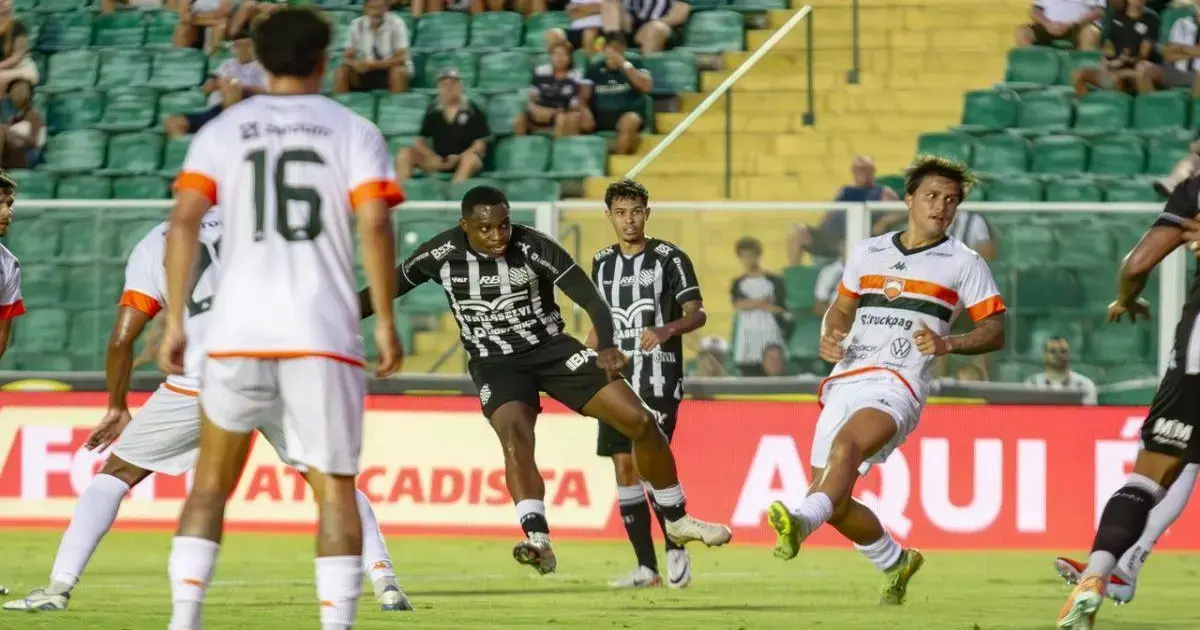 Jogadores em campo durante partida de futebol, um atleta chuta a bola enquanto outros se posicionam.