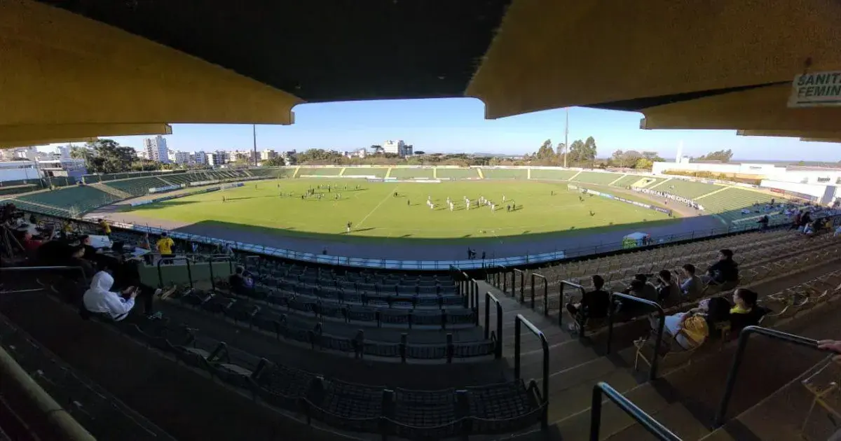 Estádio vazio com campo gramado, torcedores nas arquibancadas e jogadores em aquecimento.