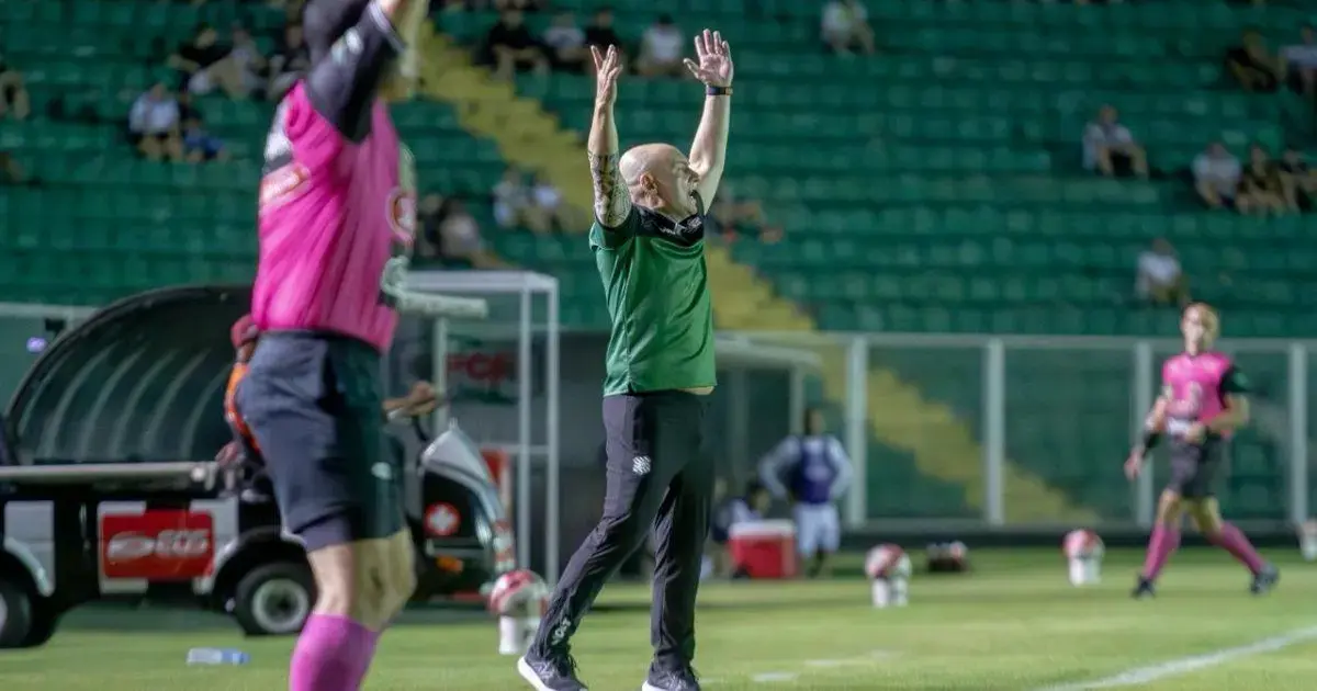 Técnico gesticula em campo, com mãos levantadas, enquanto árbitro observa ao fundo em estádio vazio.
