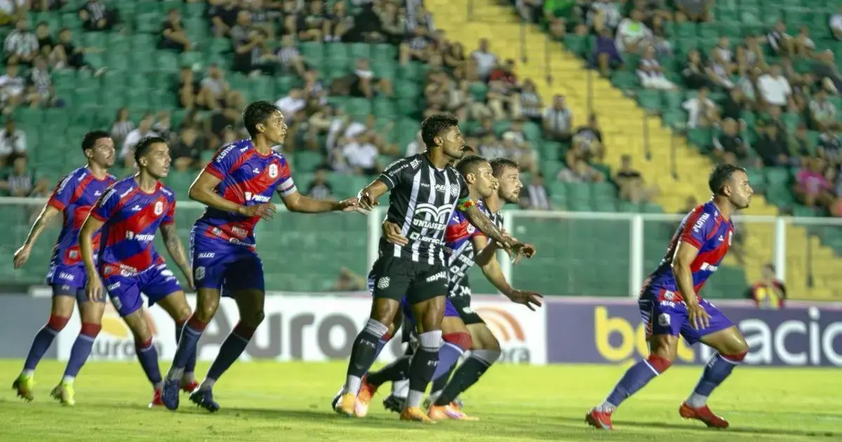 Jogadores em campo durante partida de futebol entre Figueirense e Marcílio Dias, com torcida ao fundo.