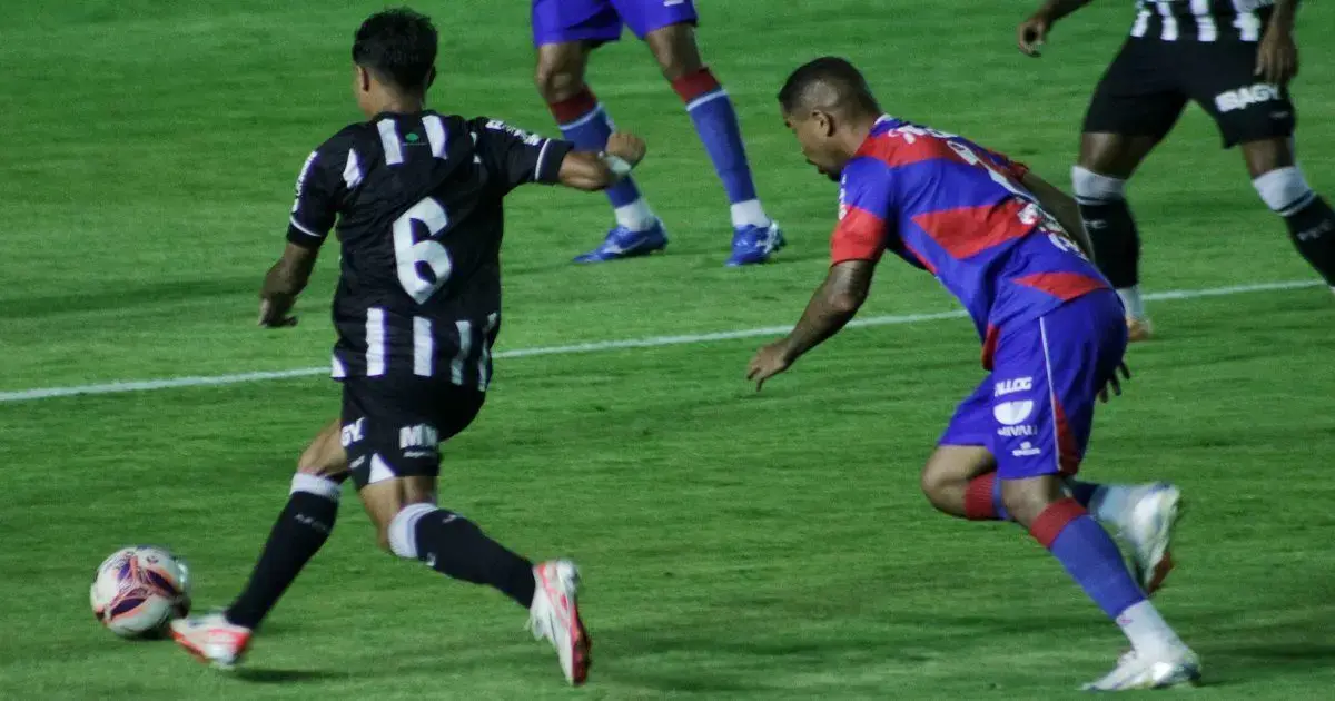 Jogadores em campo, um com uniforme preto e branco, outro com uniforme azul e vermelho, disputando a bola.