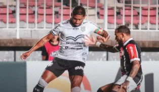 Jogadores em campo, um em uniforme branco driblando o adversário com uniforme preto e vermelho.
