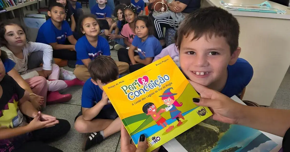 Crianças em sala de aula com camisetas azuis, uma delas segurando livro colorido sobre educação.