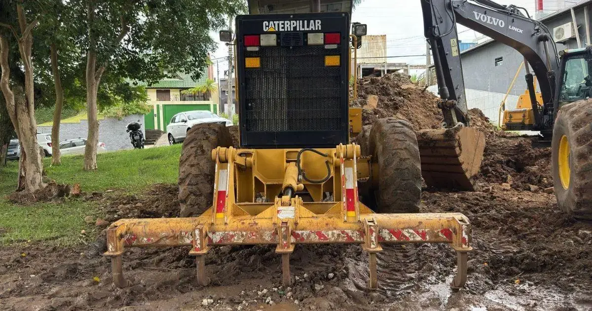 Máquina de construção em área de terra batida, com vegetação ao fundo e equipamentos pesados ao lado.