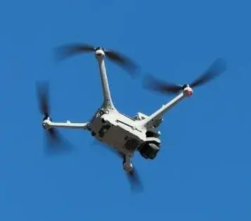 Drone autônomo voando em céu azul, com hélices visíveis e corpo branco.