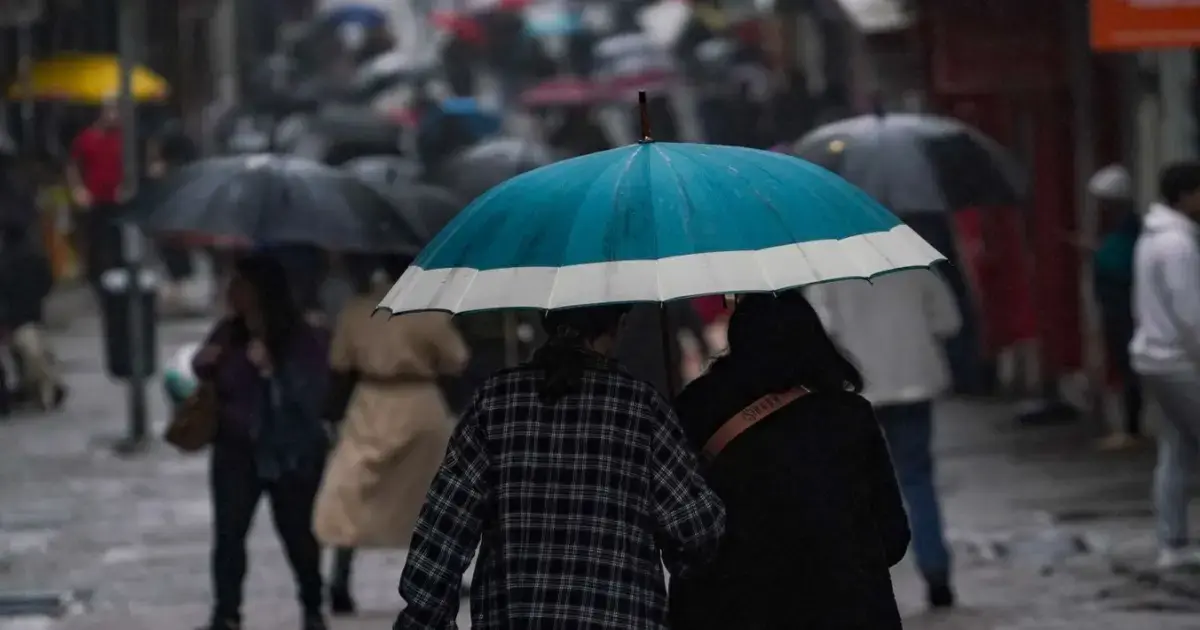 Duas pessoas caminhando sob um guarda-chuva azul em uma rua movimentada com chuva e outras pessoas ao fundo.