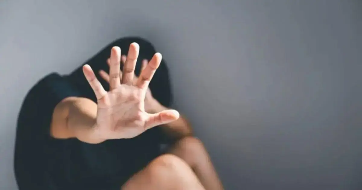 Mão estendida em gesto de defesa, mulher agachada em posição de vulnerabilidade, fundo neutro.